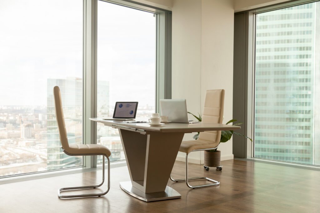 Image of work desk with two laptops on it and comfortable armchairs beside in bright office interior with large windows and urban landscape outside. Modern office workplace, negotiation room design